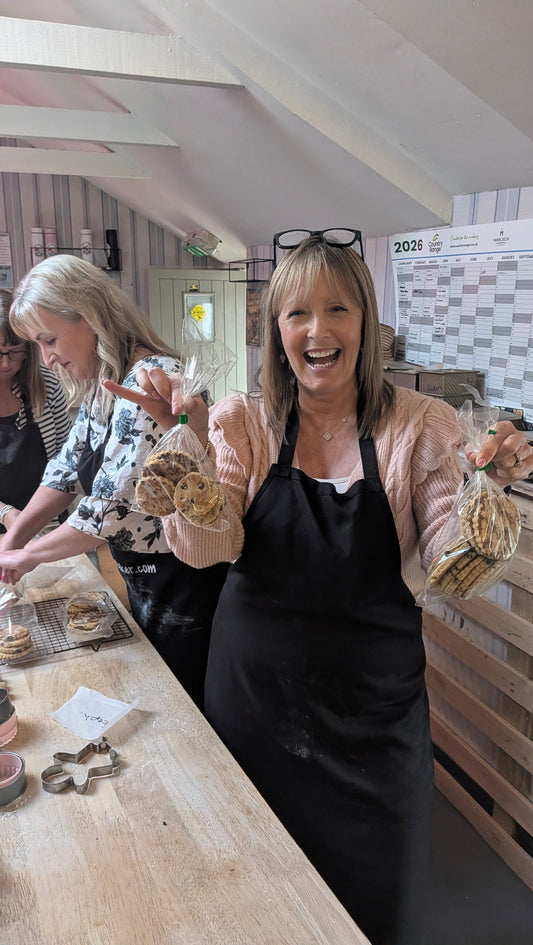 A woman in a black apron smiles while holding packaged cookies in both hands, standing at a wooden table in a kitchen or bakery setting. Another woman is visible in the background working at the counter. They are the studio of Jamie the Baker, Carmarthenshire. Llansteffan