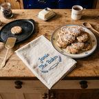 A rustic kitchen scene with a wooden countertop holding a plate of powdered pastries, a black skillet with a small pastry, a cloth napkin embroidered 'Jamie The Baker', mugs, a butter dish, and wooden utensils.